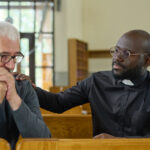 black man in shirt with clerical collar keeping hand on shoulder of parishioner